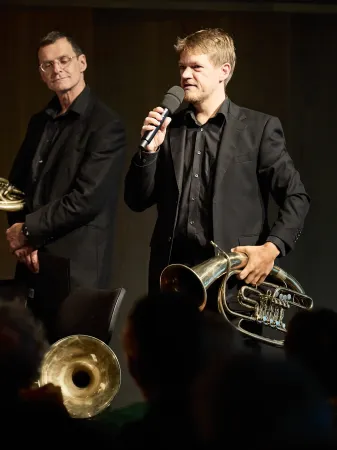 © Bregenzer Festspiele / Anja Koehler Ein Musiker in schwarzem Anzug hält ein Mikrofon in der rechten Hand und eine Tuba in der linken Hand. Im Hintergrund steht ein weiterer Musiker mit einem Horn.
