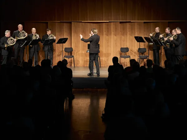 © Bregenzer Festspiele / Anja Koehler Ein Orchester mit sieben Hornspielern in schwarzer Kleidung steht auf einer Bühne. Ein Dirigent in einem schwarzen Anzug leitet sie. Im Hintergrund sind Holzpaneele sichtbar.