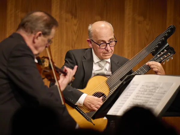 © Bregenzer Festspiele / Anja Koehler Zwei Musiker auf einer Bühne. Der Mann links spielt eine Violine, der Mann rechts spielt eine Gitarre mit sechs Saiten. Notenblätter sind auf einem Ständer sichtbar.