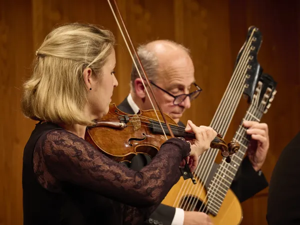 © Bregenzer Festspiele / Anja Koehler Eine Frau mit blonden Haaren spielt eine Geige. Im Hintergrund steht ein Mann mit einer Gitarre und einem großen Saiteninstrument.