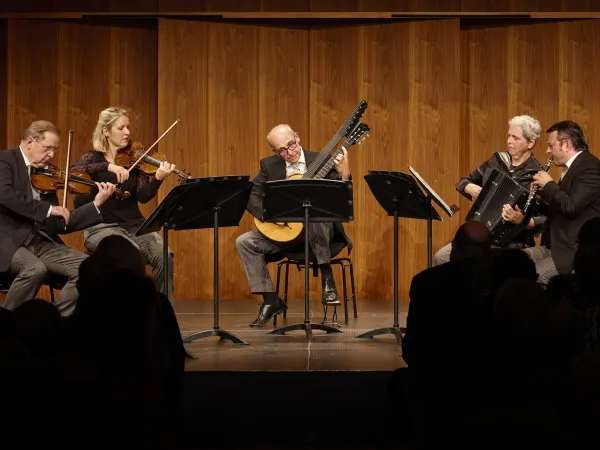 © Bregenzer Festspiele / Anja Koehler Fünf Musiker auf einer Bühne, die verschiedene Instrumente spielen: zwei Geigen, ein Akkordeon und eine Gitarre. Sie tragen formelle Kleidung und stehen um Notenständer.