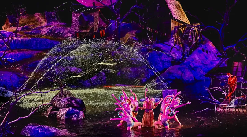 Bühnenbild von 'Der Freischütz' mit Tänzern in farbenfrohen Kostümen vor einem großen, leuchtenden Vollmond und einer mystischen Waldkulisse