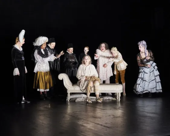 Szenenbild mit mehreren Personen in historischen Kostümen, eine Person sitzt auf einer Chaiselongue, andere stehen um sie herum auf einer dunklen Bühne.