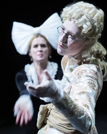 Zwei Personen in historischen Kostümen, eine mit weißer Perücke und besticktem Jackett, die andere mit großem weißem Haarschmuck, beide strecken die Hände aus.