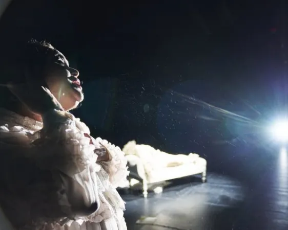 Frau in weißer, rüschiger Bluse hält sich den Kopf, im Hintergrund ein helles Sofa und ein starker Lichtschein auf dunkler Bühne.