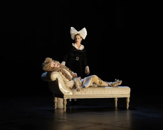 Person in schwarzem Kleid mit großer weißer Schleife steht neben liegender Person in barockem Kostüm auf einer antiken Chaiselongue.