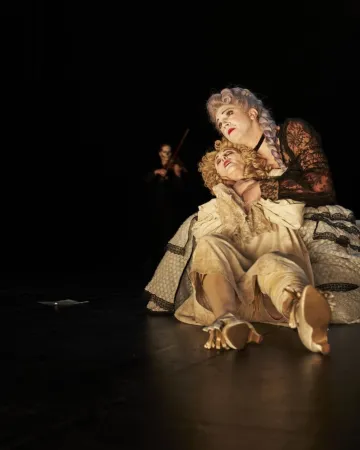 Zwei Personen in historischen Kostümen, eine sitzt am Boden, die andere hält sie im Arm, im Hintergrund eine Person mit Geige.