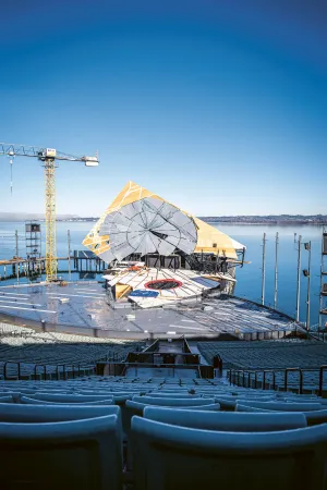 An open-air stage with rows of seats in front of a stage featuring a large, circular set piece and a crane by the lakeside under a clear sky.