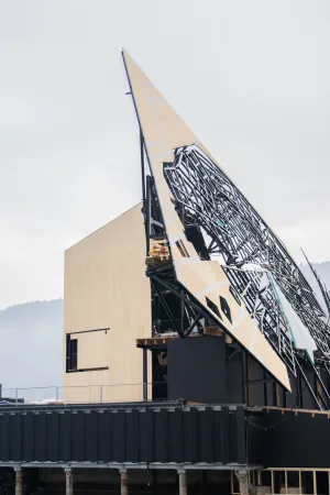 Große Bühnenkulisse mit spitz zulaufender, teilweise mit schwarzem Muster versehener Fläche und rechteckigem Holzaufbau im Hintergrund