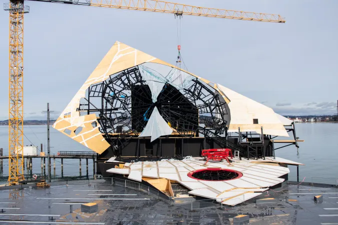 Bühnenaufbau mit großem, dreieckigem Bühnenbild und schwarzer Stahlkonstruktion auf einer Wasserbühne mit Kran im Hintergrund
