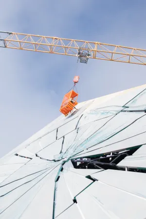 Kran hebt orangefarbenen Lastkorb über eine große, zerbrochene Glasfläche mit sichtbaren Rissen und Bruchstellen