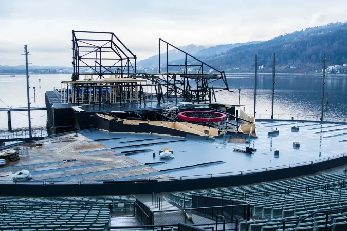 Bühnenaufbau mit Stahlgerüst und runden Elementen vor einem See und bewaldeten Hügeln im Hintergrund
