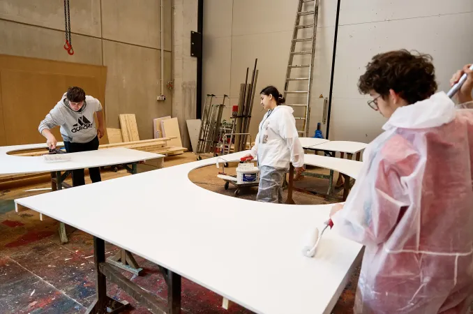 Drei Personen in Schutzkleidung streichen große weiße Holzplatten in einer Werkstatt mit Farbrollern und Pinsel