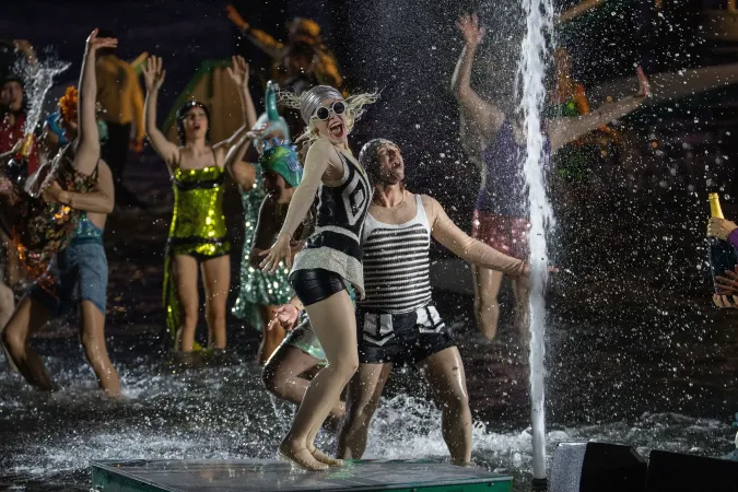 © Bregenzer Festspiele / Daniel Ammann Menschen in bunten Kostümen tanzen und springen um eine Wasserfontäne auf einer Bühne