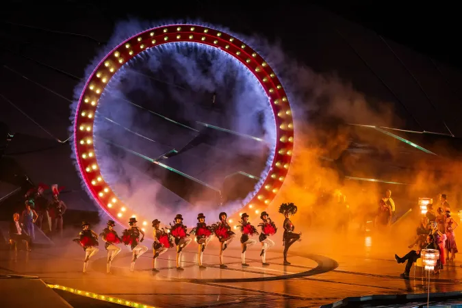 © Bregenzer Festspiele / Daniel Ammann Tänzerinnen in roten Kostümen mit schwarzen Federkopfschmuck führen eine Choreografie vor einem großen, beleuchteten Ring mit Nebel auf einer Bühne auf