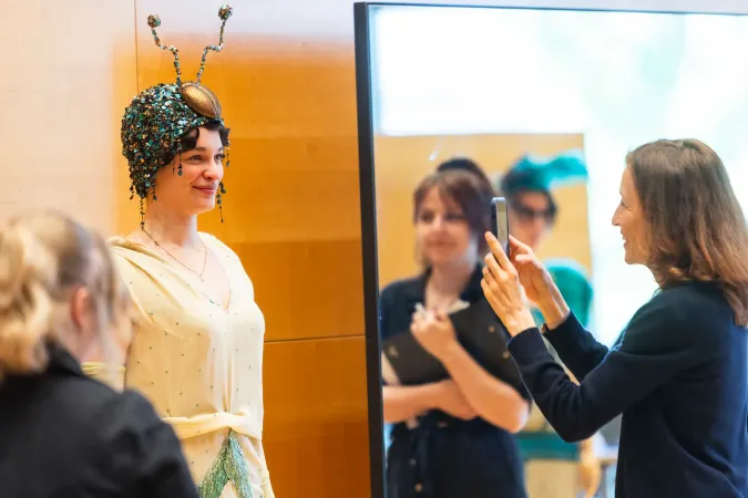 © Bregenzer Festspiele / Kristina Becker Frau mit fantasievollem Kopfschmuck in Form von Insektenantennen steht vor Spiegel, während eine andere Frau sie mit Smartphone fotografiert