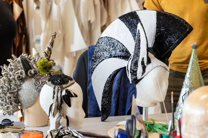 © Bregenzer Festspiele / Kristina Becker Drei fantasievolle Kostümkopfbedeckungen auf weißen Schaufensterpuppenköpfen auf einem Tisch, darunter ein Einhorn, ein schwarz-weiß gestreifter Helm und eine glitzernde grüne Zipfelmütze
