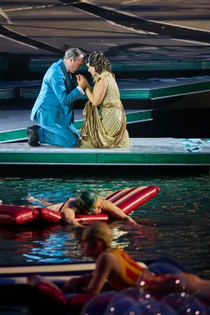 © Bregenzer Festspiele / Anja Koehler Ein Mann im blauen Anzug und eine Frau im goldenen Kleid knien sich auf einer Bühne über Wasser und halten sich an den Händen, im Vordergrund liegen zwei Personen auf roten Luftmatratzen im Wasser.