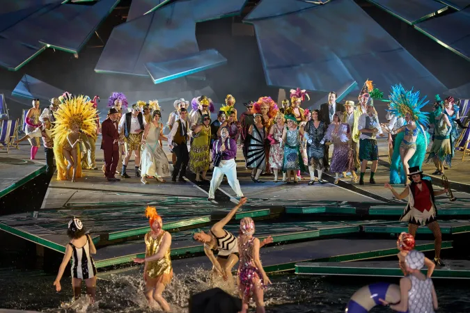 © Bregenzer Festspiele / Anja Koehler Bühnenensemble in farbenfrohen Kostümen mit Federschmuck und Badebekleidung auf einer Bühne mit Wasserbecken