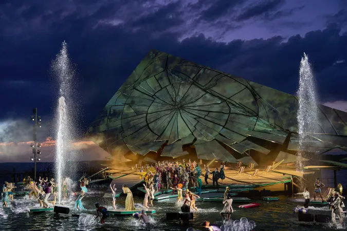 © Bregenzer Festspiele / Anja Koehler Bühnenbild mit großem schräg gestelltem, zerbrochen wirkendem Rad im Hintergrund und zahlreichen Schauspielern auf einer Wasserbühne mit zwei hohen Wasserfontänen bei Dämmerung