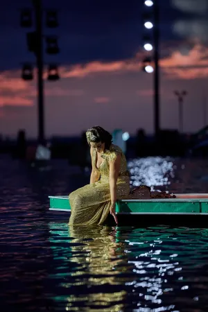 © Bregenzer Festspiele / Anja Koehler Frau in langem, glitzerndem Kleid sitzt am Rand eines Stegs und berührt mit der Hand das Wasser bei Dämmerung