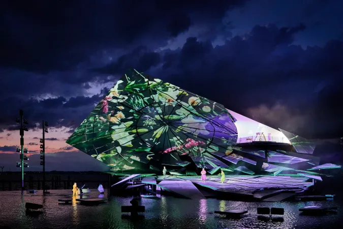 © Bregenzer Festspiele / Anja Koehler Bühnenaufbau mit farbiger Projektion auf einer Wasserbühne unter bewölktem Abendhimmel mit mehreren Personen auf der Bühne und im Wasser