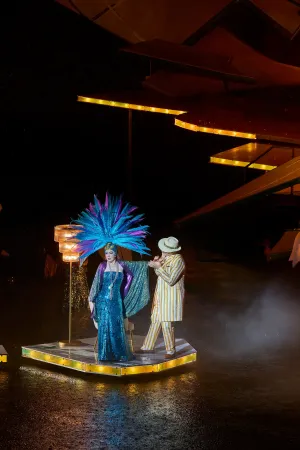 © Bregenzer Festspiele / Anja Koehler Zwei Personen auf einer beleuchteten Bühne, eine trägt ein blaues Paillettenkleid und einen großen blauen Federkopfschmuck, die andere einen gestreiften Anzug und einen Hut