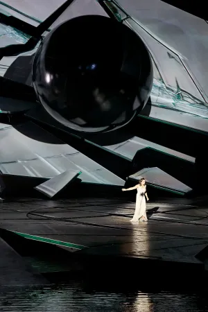 © Bregenzer Festspiele / Anja Koehler Eine Frau in langem, hellem Kleid steht auf einer Bühne vor einer großen, abstrakten, schwarzen und weißen Bühnenkulisse mit geometrischen Formen und einer großen schwarzen Kugel