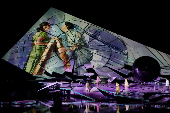 © Bregenzer Festspiele / Anja Koehler Bühnenbild mit großer gebrochener Spiegelfläche, auf der zwei Frauen in historischen Kostümen zu sehen sind, davor mehrere Personen auf einer abstrakten Bühne über Wasser