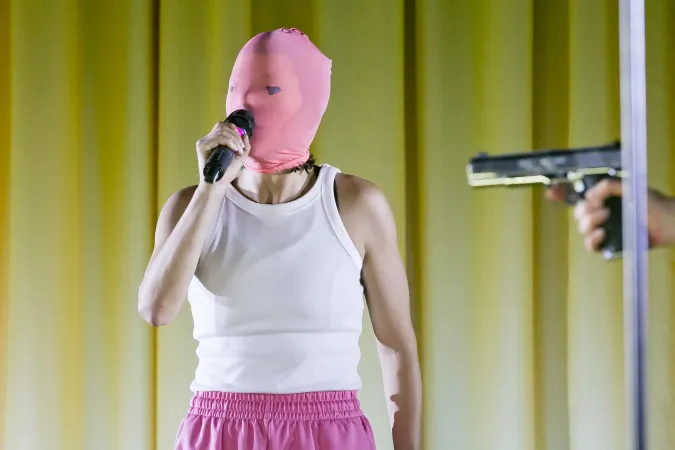 Person mit rosa Maske und weißem Tanktop hält Mikrofon vor gelbem Hintergrund, im Vordergrund ist eine Pistole zu sehen.