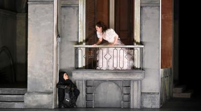 Tancredi sitzend vor einem Balkon, Amenaide im Hochzeitskleid beugt sich zu ihr. 